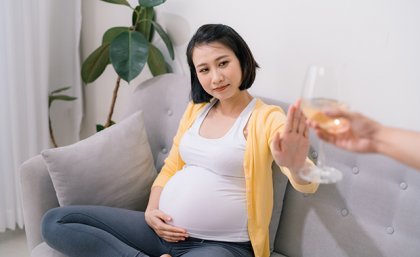 A pregnant woman sitting on a couch refusing an offered glass of wine